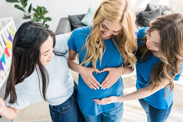 Vista de ángulo alto de amigos multiculturales y mujeres embarazadas tocando el vientre y haciendo corazón con las manos en la fiesta del bebé - foto de stock