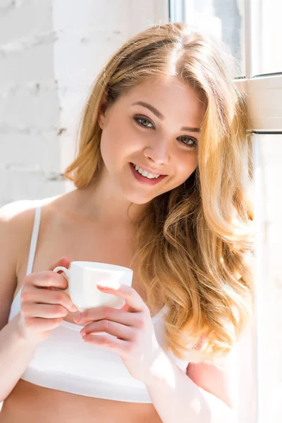 Beautiful cheerful girl with cup of coffee standing near window — Stock Photo