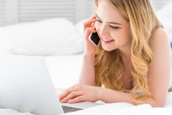 Menina sorrindo falando no smartphone e usando laptop na cama — Fotografia de Stock