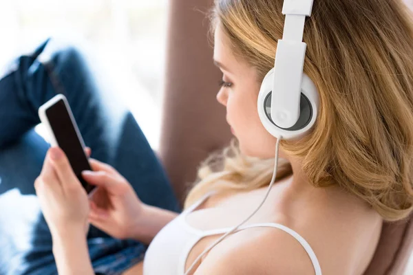 Blonde girl using smartphone and listening music with headphones on bed — Stock Photo