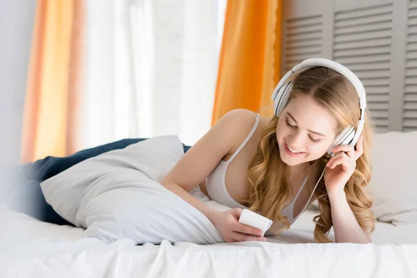 Cheerful girl using smartphone and listening music with headphones while lying on bed — Stock Photo