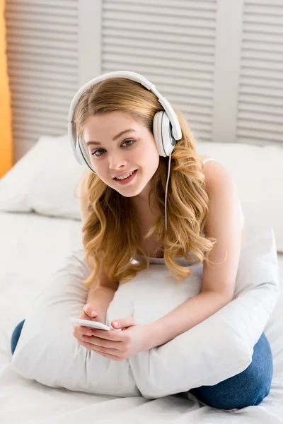 Дівчина використовує смартфон і слухає музику з навушниками, сидячи на ліжку — стокове фото