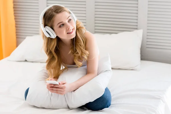 Chica sonriente usando teléfono inteligente y escuchar música con auriculares en la cama - foto de stock