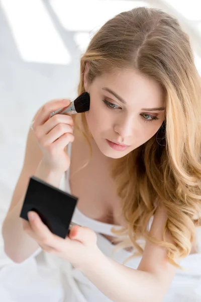 Menina atraente aplicando blush com escova e olhando para o espelho — Fotografia de Stock