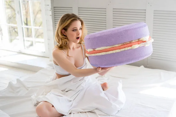 Menina surpreso segurando grande macaron violeta na cama — Fotografia de Stock