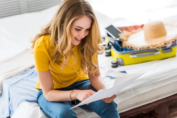 Attractive girl looking at map while sitting on bed with suitcase — Stock Photo