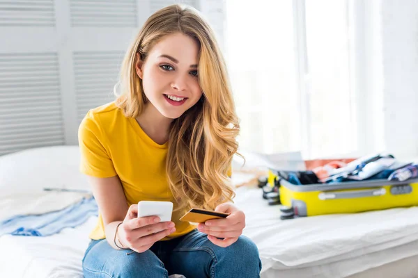 Menina sorrindo usando smartphone e cartão de crédito com saco de viagem na cama — Fotografia de Stock