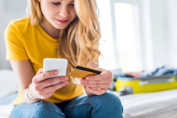 Hermosa chica usando teléfono inteligente y tarjeta de crédito con maleta en la cama - foto de stock