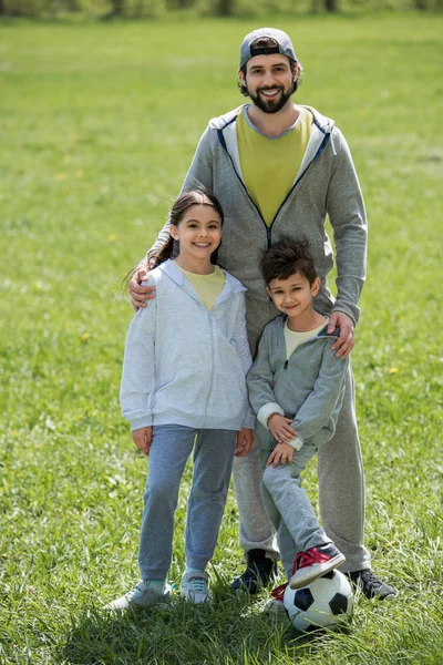 Sonriente padre sosteniendo hija e hijo con pelota de fútbol en el parque - foto de stock