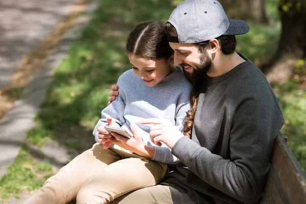 Felice padre e figlia utilizzando smartphone su panchina nel parco — Foto stock