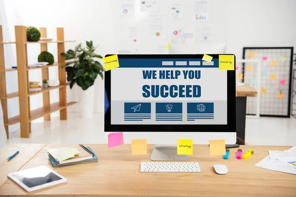 Close up view of workplace with we help you succeed inscription on computer screen and sticky notes in office — Stock Photo
