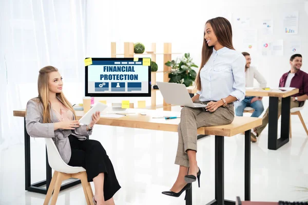 Selektiver Fokus multiethnischer Geschäftsfrauen am Arbeitsplatz mit finanzieller Schutzbeschriftung auf dem Bildschirm und Kollegen im Büro dahinter — Stockfoto
