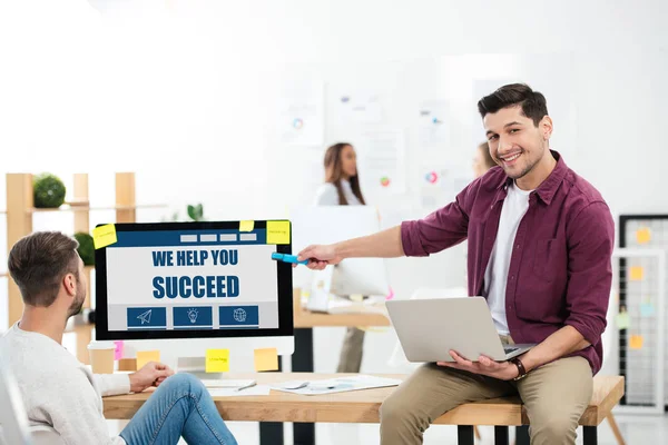 Businessman pointing at we help you succeed inscription on computer screen while working at workplace with colleague in office — Stock Photo