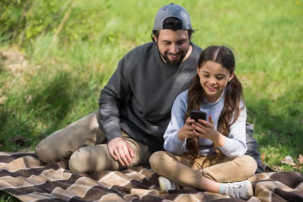 Smiling father watching daughter using smartphone on plaid in park — Stock Photo
