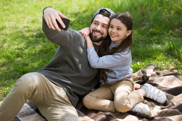 Smiling father taking selfie with daughter on plaid in park — Stock Photo