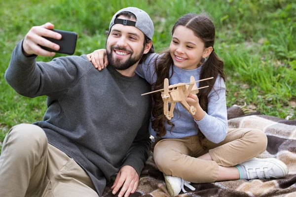 Padre scattare selfie con figlia in possesso di aereo giocattolo in legno sul plaid nel parco — Foto stock