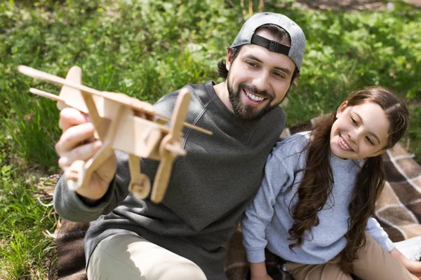 Sonriente padre e hija jugando con juguete avión de madera en cuadros en el parque - foto de stock