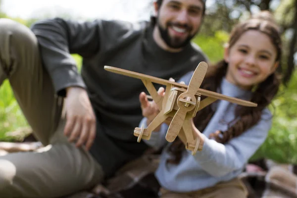 Vue rapprochée de jouet en bois avion dans les mains de la fille assise sur plaid avec père dans le parc — Photo de stock