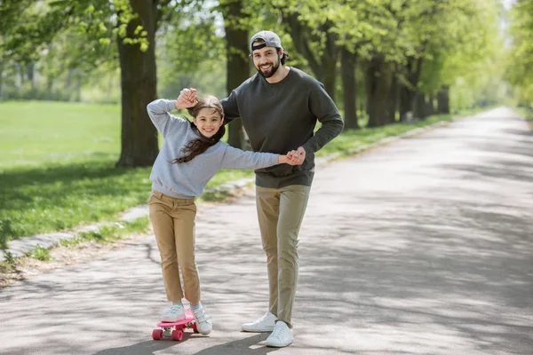 Sonriente padre ayudando a su hija a montar en monopatín en el parque - foto de stock