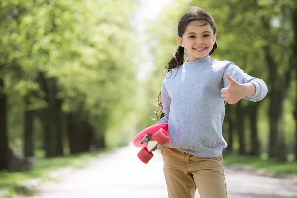 Niño sonriente con monopatín haciendo gesto de pulgar hacia arriba en el parque - foto de stock