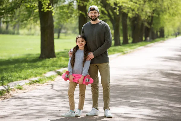 Sonriente padre e hija sosteniendo monopatín en el parque - foto de stock