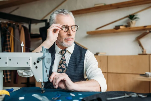 Seniorschneiderin sitzt mit Nähmaschine am Tisch und schaut in Nähwerkstatt — Stockfoto