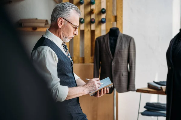 Tailleur senior écrivant quelque chose pour ordinateur portable à l'atelier de couture — Photo de stock