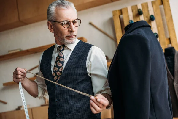 Senior tailor holding tape measure and looking at jacket at sewing workshop — Stock Photo