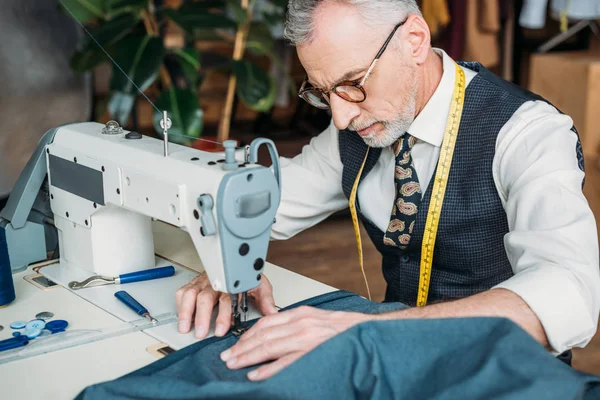 Handsome tailor in glasses sewing cloth with sewing machine at sewing workshop — Stock Photo