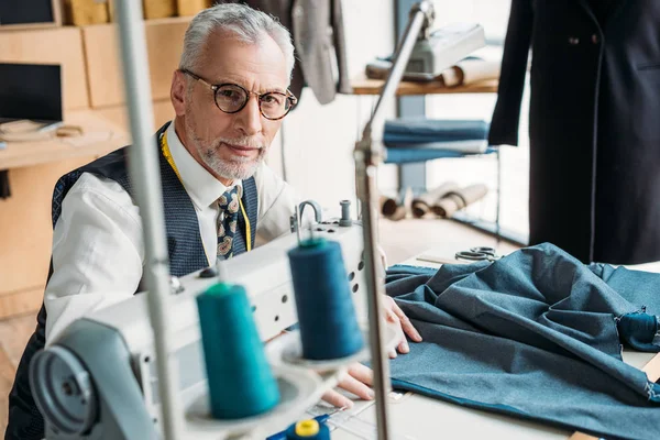 Handsome tailor sewing cloth with sewing machine and looking at camera at sewing workshop — Stock Photo