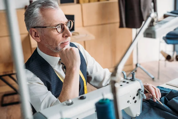 Pensive tailor sitting at table with sewing machine and looking away at sewing workshop — Stock Photo