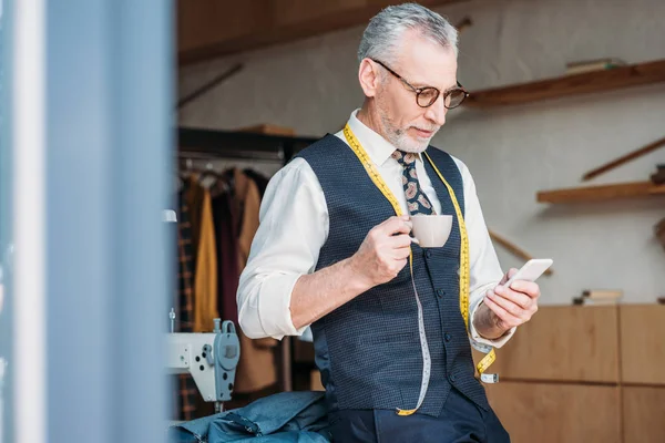 Handsome tailor standing with cup of coffee and looking at smartphone at sewing workshop — Stock Photo