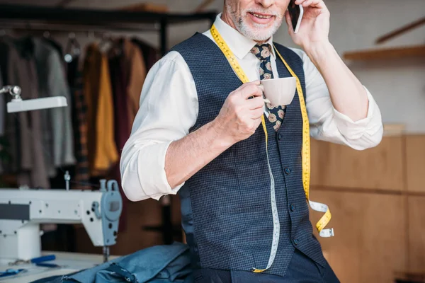 Abgeschnittenes Bild eines Schneiders, der mit Tasse Kaffee in der Nähwerkstatt steht und mit dem Smartphone spricht — Stockfoto