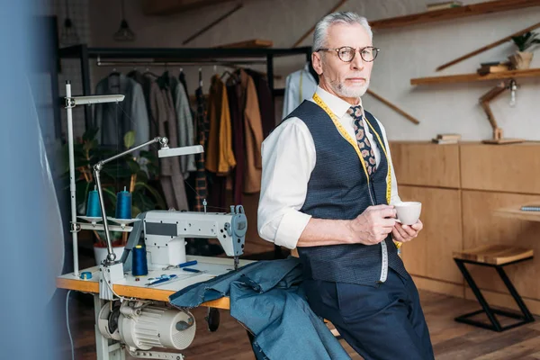 Schöne Schneiderin steht mit Tasse Kaffee in Nähwerkstatt — Stockfoto