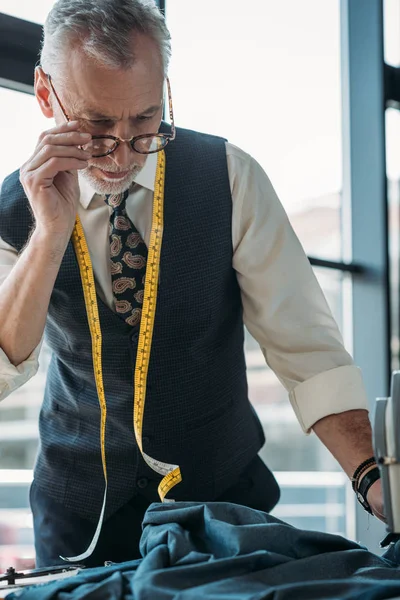 Schöne Schneiderin berührt Brille und schaut Stoff in Nähwerkstatt an — Stockfoto