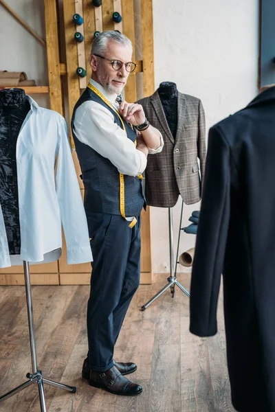Guapo sastre de pelo gris mirando chaquetas en maniquíes en taller de costura - foto de stock