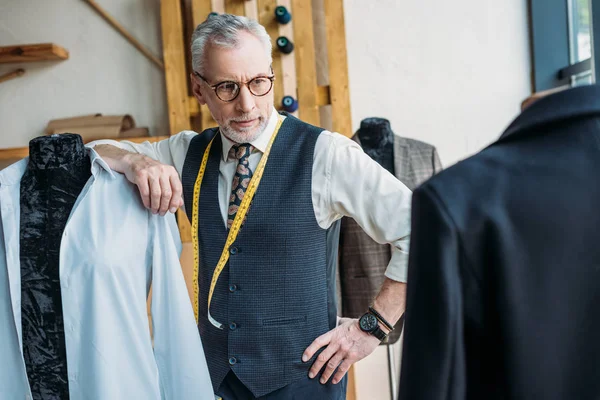 Handsome tailor leaning on mannequin at sewing workshop and looking at jacket — Stock Photo