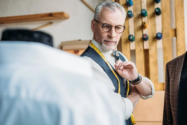 Bello sarto anziano guardando giacche su manichini a laboratorio di cucito — Foto stock