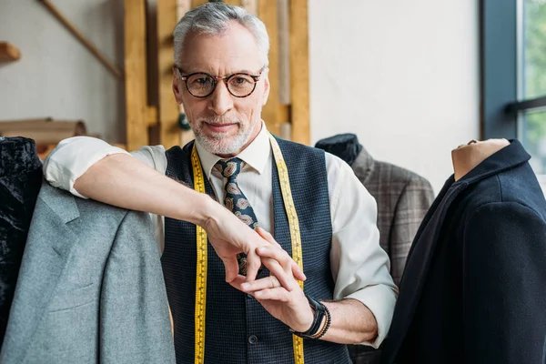 Tailleur mature souriant avec ruban à mesurer appuyé sur le mannequin avec veste à l'atelier de couture et regardant la caméra — Photo de stock