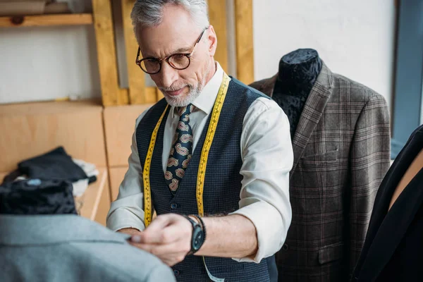Handsome mature tailor with measuring tape looking at jacket on mannequin at sewing workshop — Stock Photo
