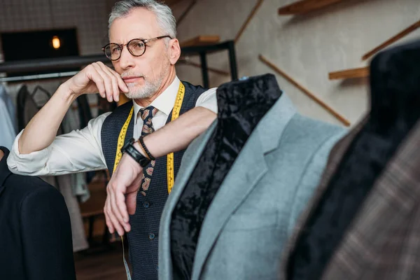Thoughtful mature tailor with measuring tape leaning on mannequin with jacket at sewing workshop — Stock Photo
