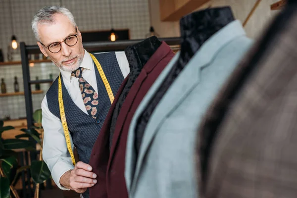 Tailleur mature concentré avec ruban à mesurer et bloc-notes examinant les vestes sur mannequins à l'atelier de couture — Photo de stock