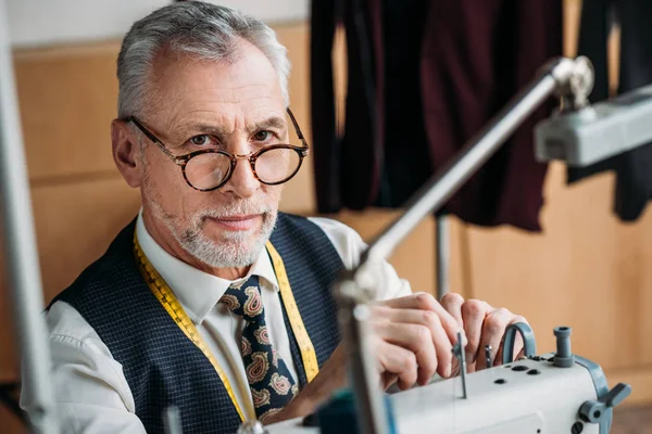 Elegante alfaiate maduro trabalhando com máquina de costura na oficina e olhando para a câmera — Fotografia de Stock
