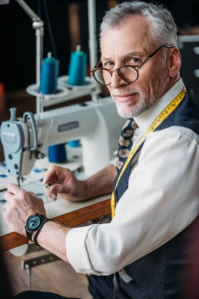 Stylish mature tailor working with sewing machine at workshop and looking at camera — Stock Photo