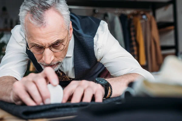 Close-up shot of handsome mature tailor marking cloth pattern with chalk at sewing workshop — Stock Photo