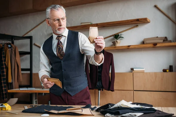 Stilvolle reife Schneiderin schaut sich Papiertaschenmuster an, um es in der Nähwerkstatt auf Stoff zu verschieben — Stockfoto