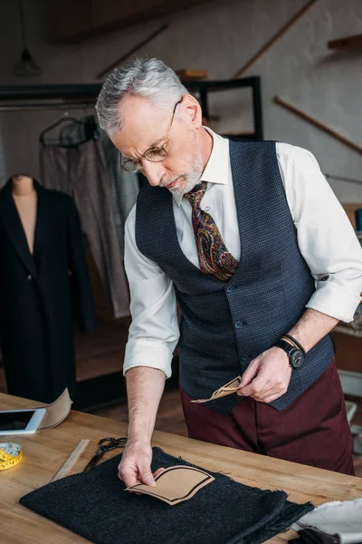 Serious mature tailor looking at paper pockets pattern to postpone it on cloth at sewing workshop — Stock Photo