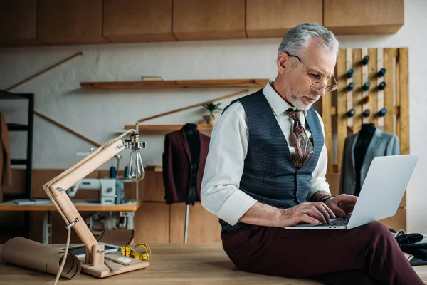 Tailleur mature concentré travaillant avec ordinateur portable tout en étant assis sur la table à l'atelier de couture — Photo de stock