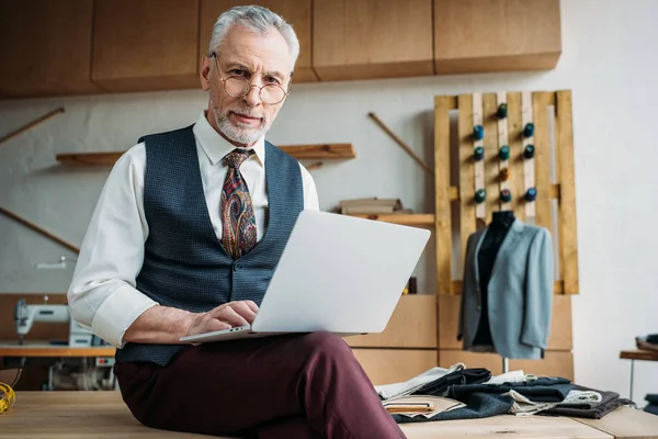 Elegante sarto maturo che lavora con il computer portatile mentre seduto sul tavolo al laboratorio di cucito — Foto stock