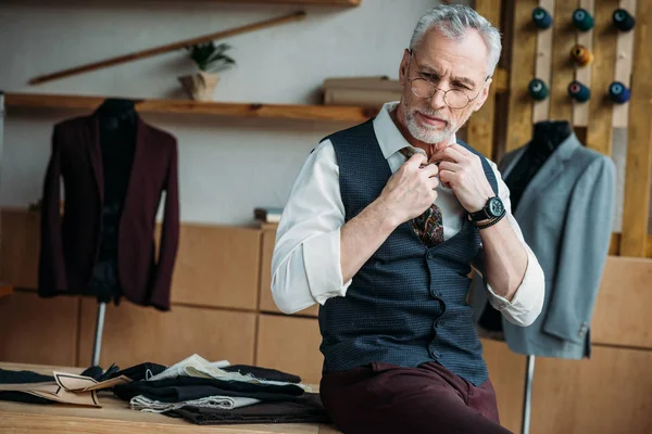 Handsome mature tailor in stylish clothing sitting on work desk at sewing workshop — Stock Photo